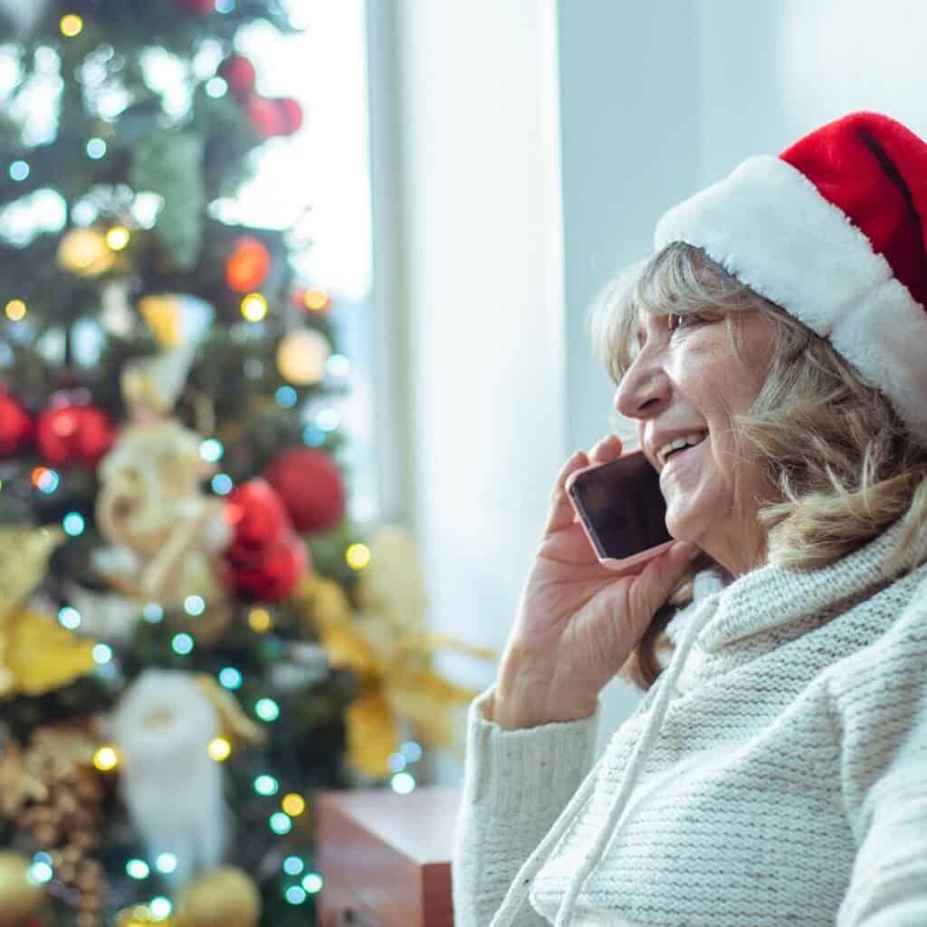 Older woman on a cell phone wearing a santa hat with a decorated tree in the background