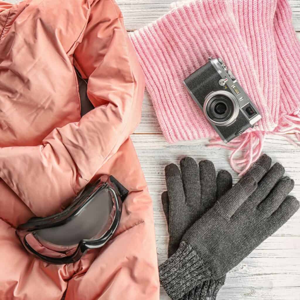 Winter gear in grey, pink and peach tones laid out with an old school snapshot camera on a white wood background